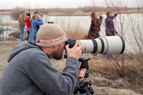 A photographer in a gray hoodie captures a scene with a large camera, while others observe in the background by the water.