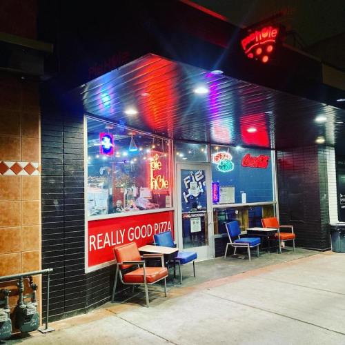 A brightly lit pizza shop entrance with colorful signage and outdoor seating at night.