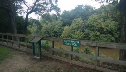 A wooden railing with a sign reading "Bayou Macon Overlook" beside a calm river and lush green trees.