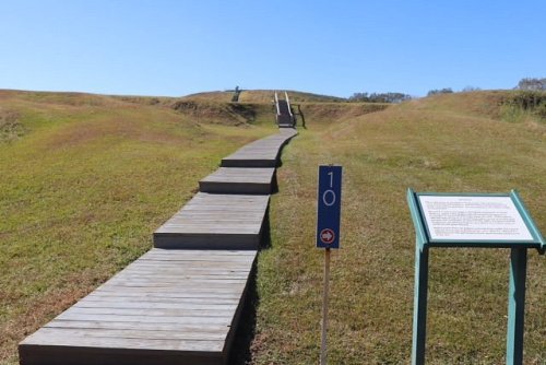 A wooden walkway leads up a grassy hill, with a signpost and informational board nearby under a clear blue sky.
