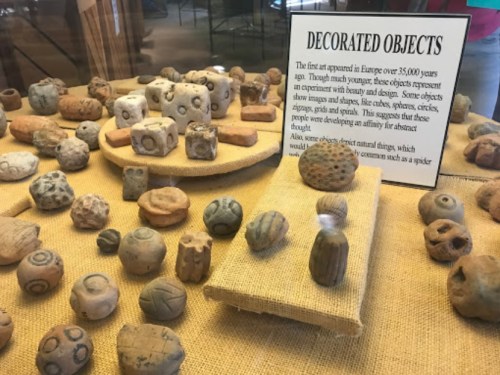 A display of ancient decorated objects, including various shapes and designs, on a textured surface in a museum setting.