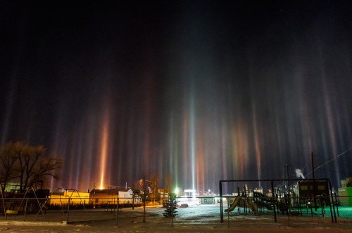 Colorful light pillars rise into the night sky above a snowy playground, illuminating a quiet town scene.