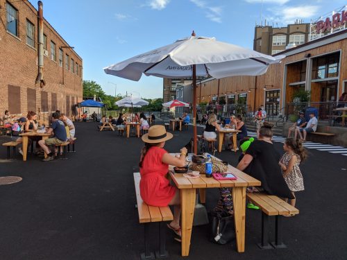 Outdoor dining scene with people at wooden tables under umbrellas, enjoying food and drinks in a lively atmosphere.