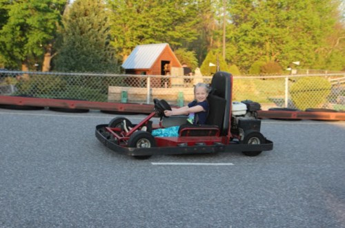 A child smiles while driving a go-kart on a racetrack surrounded by trees and a fence.
