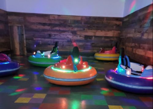 Colorful bumper cars with lights are arranged in a playful indoor space with a wooden wall and patterned floor.