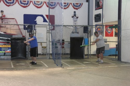 Two men practice batting in a baseball cage, surrounded by nets and sports-themed decor.