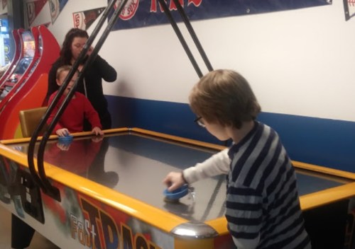 A child plays air hockey while a woman and another child watch, in an arcade setting.