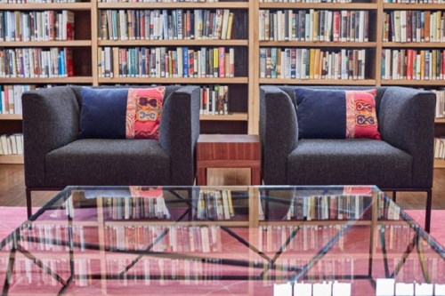 Two cozy chairs with decorative pillows face a glass coffee table, surrounded by shelves of books in a library setting.