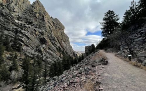 A rocky trail winds through a canyon, flanked by towering cliffs and scattered pine trees under a cloudy sky.