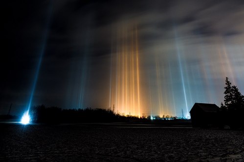 A night scene with colorful light pillars rising from the horizon, illuminating a dark landscape and a silhouette of a barn.