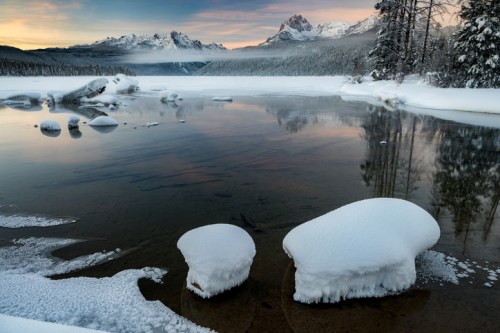 A serene winter landscape featuring a snowy shoreline, calm water, and distant mountains under a colorful sky.