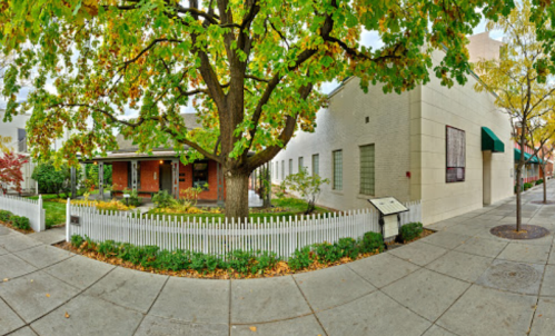 A corner view of a house with a large tree, surrounded by a white picket fence and a well-maintained garden.