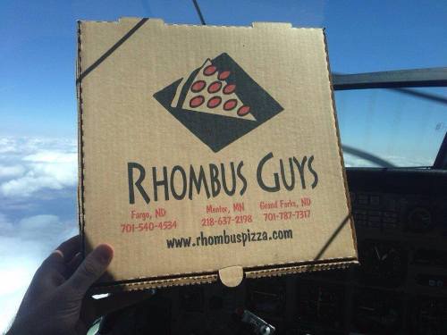 A person holds a pizza box from Rhombus Guys in an airplane cockpit, with clouds visible outside the window.