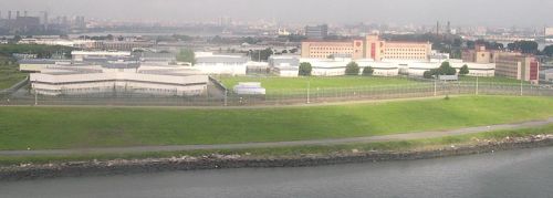 Aerial view of a large correctional facility surrounded by green grass and a river, with city buildings in the background.