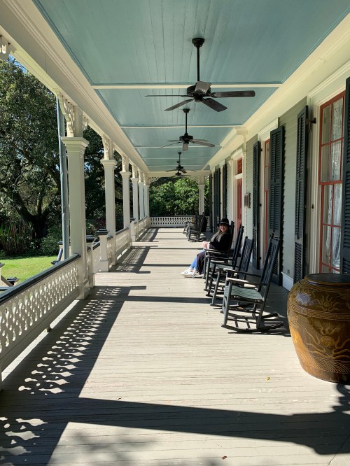A porch with rocking chairs and a person sitting, surrounded by greenery and sunlight.