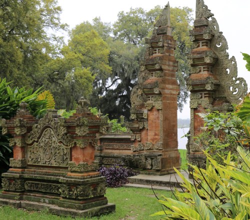 Two ornate Balinese-style stone structures surrounded by lush greenery and trees near a body of water.