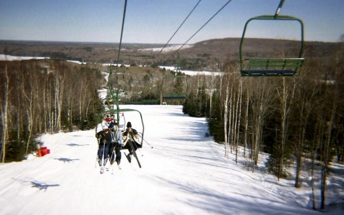 Skiers on a chairlift ascend a snowy slope, surrounded by trees and a scenic winter landscape.
