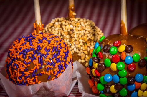 Three caramel apples decorated with colorful sprinkles and candies on a checkered tablecloth.