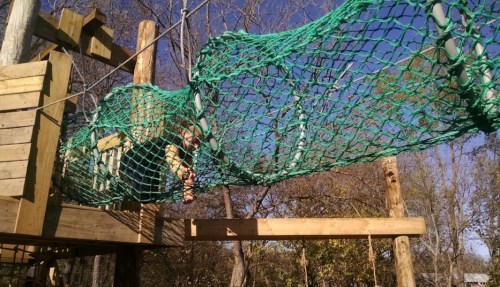 A child navigates a green rope net in a playground, surrounded by trees and a clear blue sky.