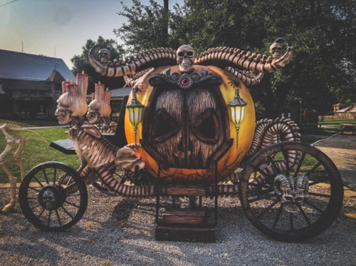 A spooky pumpkin carriage adorned with skeletons and skulls, set in a gravel area with trees in the background.