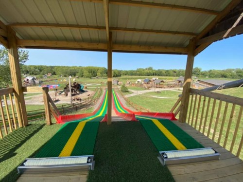View from a wooden platform overlooking colorful slides leading down to a grassy play area and playground in the distance.