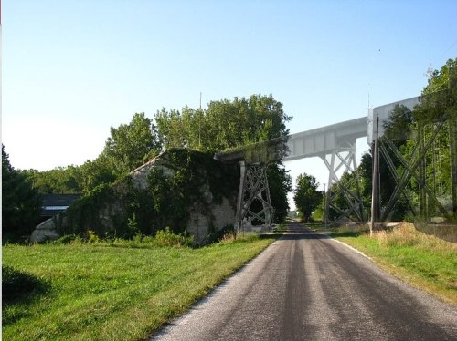 A rural road leads to an old, overgrown bridge structure surrounded by trees and greenery.