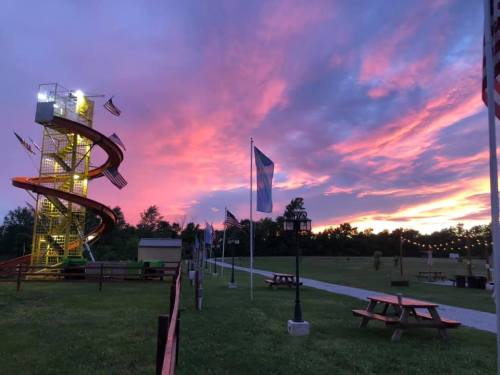 A colorful sunset over a park featuring a spiral slide, picnic tables, and flags along a pathway.