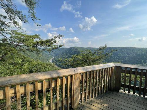 A wooden overlook with a scenic view of green hills and a winding river under a blue sky with fluffy clouds.