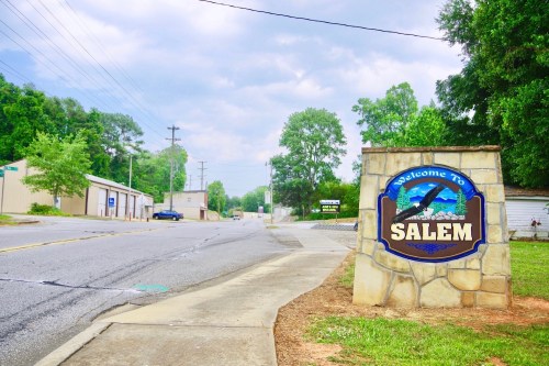 Welcome sign for Salem, with a tree-lined street and buildings in the background under a cloudy sky.