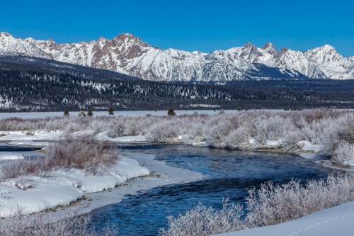 A serene winter landscape featuring a winding river, snow-covered banks, and majestic mountains under a clear blue sky.