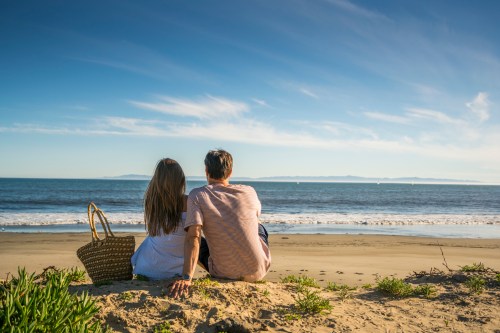 A couple sits on the beach, gazing at the ocean and distant islands under a clear blue sky.