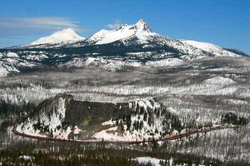 A scenic view of snow-covered mountains and a winding road through a forested landscape under a clear blue sky.