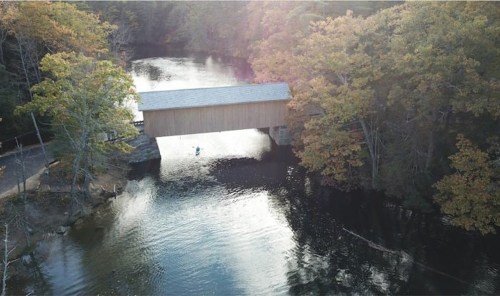 Aerial view of a covered bridge over a calm river, surrounded by autumn-colored trees.