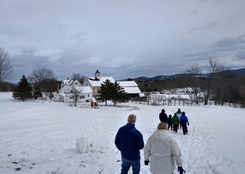 A group of people walking through snow towards a white barn and trees under a cloudy sky.