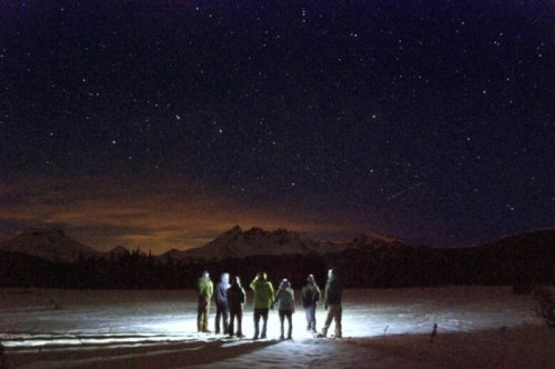 A group of people stands in a snowy landscape, illuminated by light, under a starry night sky with mountains in the background.