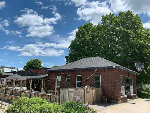 A brick building with a wooden deck, surrounded by greenery and under a blue sky with fluffy clouds.