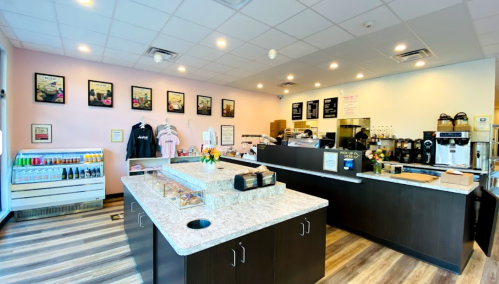 A cozy café interior with a counter, display case, and colorful drink fridge, adorned with framed pictures on the walls.