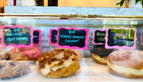 A display case of assorted donuts, labeled with flavors like Maine Blueberry, Maple Smoked Coconut, Boston Cream, and Apple Jam.