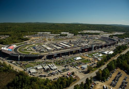 Aerial view of a large racetrack surrounded by trees and parking areas, bustling with activity and fans.