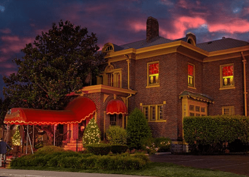 A brick house with red awnings and festive lights, set against a colorful sunset sky.