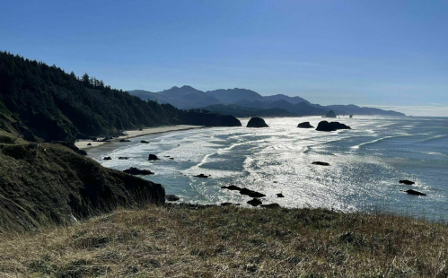 A scenic coastal view with waves, rocky formations, and mountains under a clear blue sky.