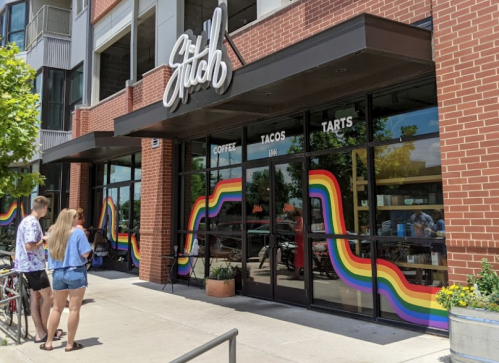 A colorful storefront with rainbow stripes, featuring "Stitch" and people waiting outside.