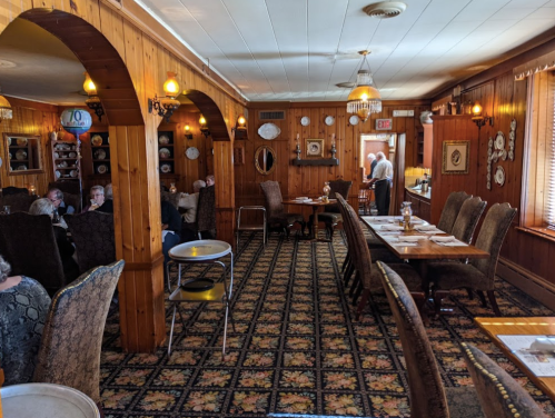 Cozy restaurant interior with wooden walls, patterned carpet, and tables set for dining. Patrons are seated and chatting.