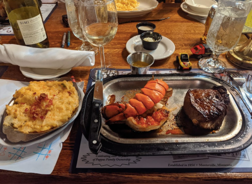 A plate featuring a lobster tail, steak, and a side of cheesy potatoes, with glasses of wine and water in the background.