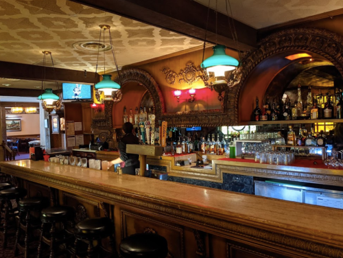 A vintage bar interior with ornate decor, a long wooden counter, and shelves stocked with various bottles.