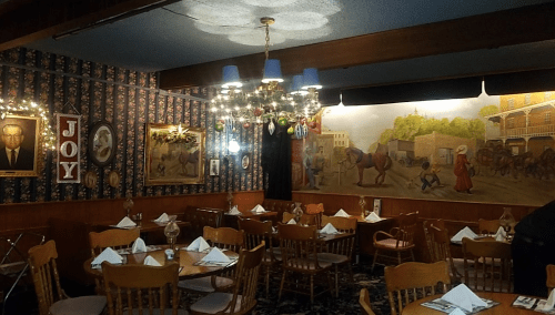 Cozy restaurant interior with wooden chairs, a chandelier, and colorful wall art depicting scenes of daily life.