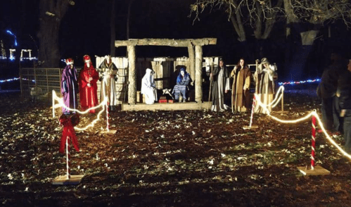 A nighttime scene of a nativity display with figures in traditional attire, surrounded by festive lights and decorations.