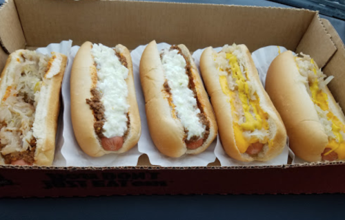 A box of five hot dogs with various toppings, including sauerkraut, chili, and cheese, on paper liners.