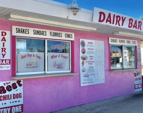 A pink dairy bar with windows displaying a menu of shakes, sundaes, and hot dogs. Bright and inviting atmosphere.