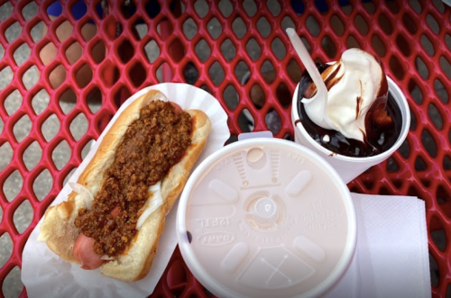 A hot dog topped with chili and a cup of soft serve ice cream with chocolate sauce on a red table.
